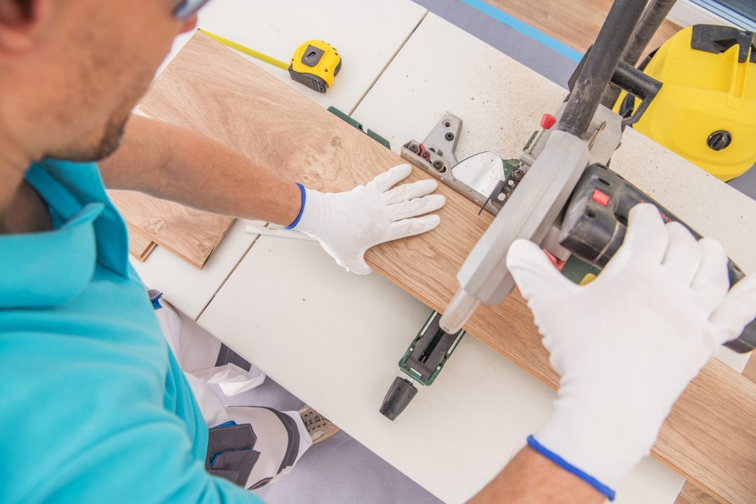 A person in gloves uses a table saw to cut a wooden board, meticulously preparing for hardwood floor installation. Measuring tape and other tools lie ready on the workbench, ensuring precision in every cut.