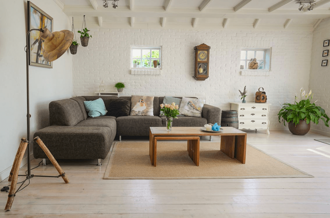 A cozy living room with a dark sectional sofa, small wooden coffee table, and white brick walls features newly completed laminate floor installation. There are decorative pillows, a wall clock, plants, and a vintage suitcase. Natural light fills the room.