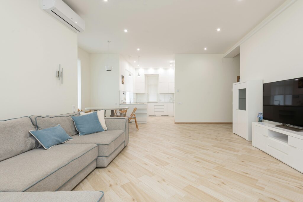 Modern living room with a gray sofa, blue cushions, flat-screen TV, and dining area. Bright lighting highlights the sleek laminate floor installation that complements the light wood accents throughout.